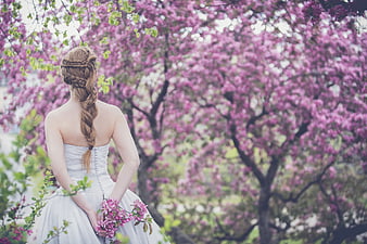 Bride in white gown with braided hair holding pink flowers in front of blooming trees