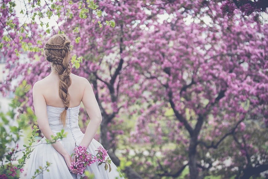 Bride in white gown with braided hair holding pink flowers in front of blooming trees