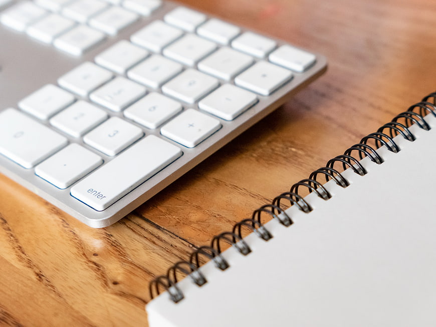 White Keyboard and Spiral Notebook on Wooden Desk