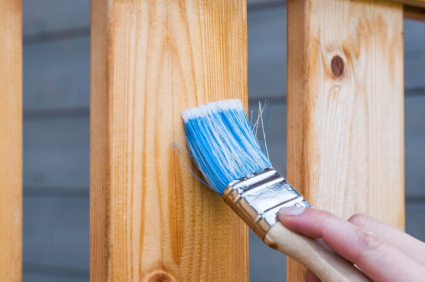 Hand applying blue paint to wooden fence with brush