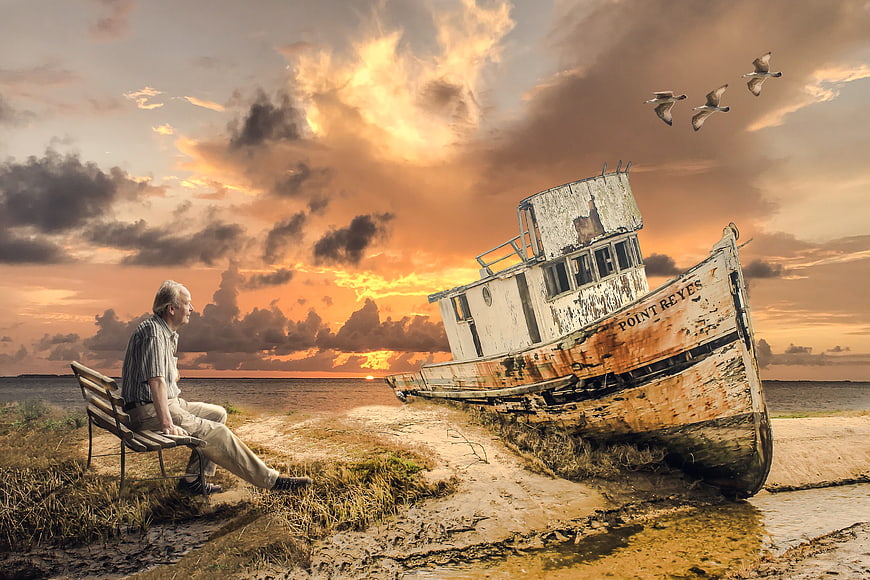 Senior man sitting on bench near old boat at sunset