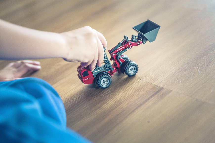 Child Playing With Red Toy Wheel Loader On Wooden Floor