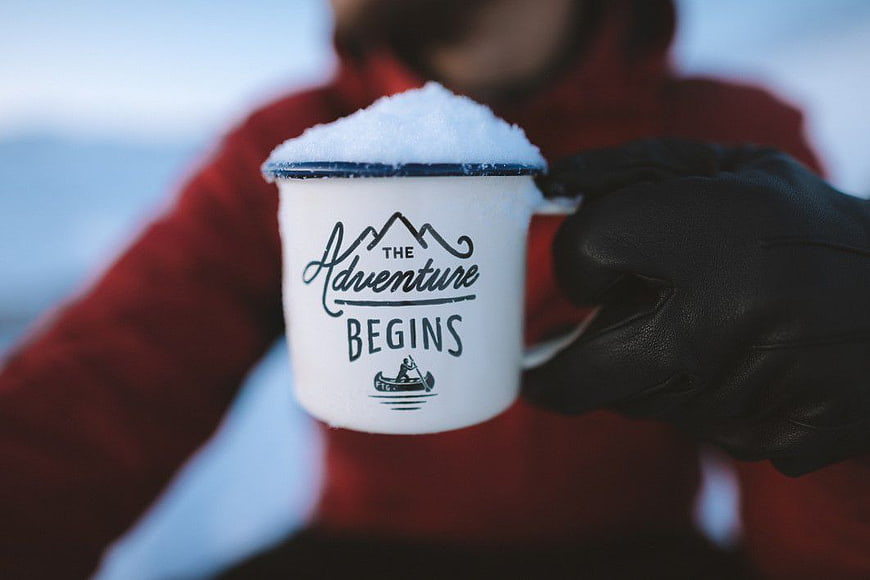 Gloved Hand Holding Snow-Filled Mug with Adventure Text