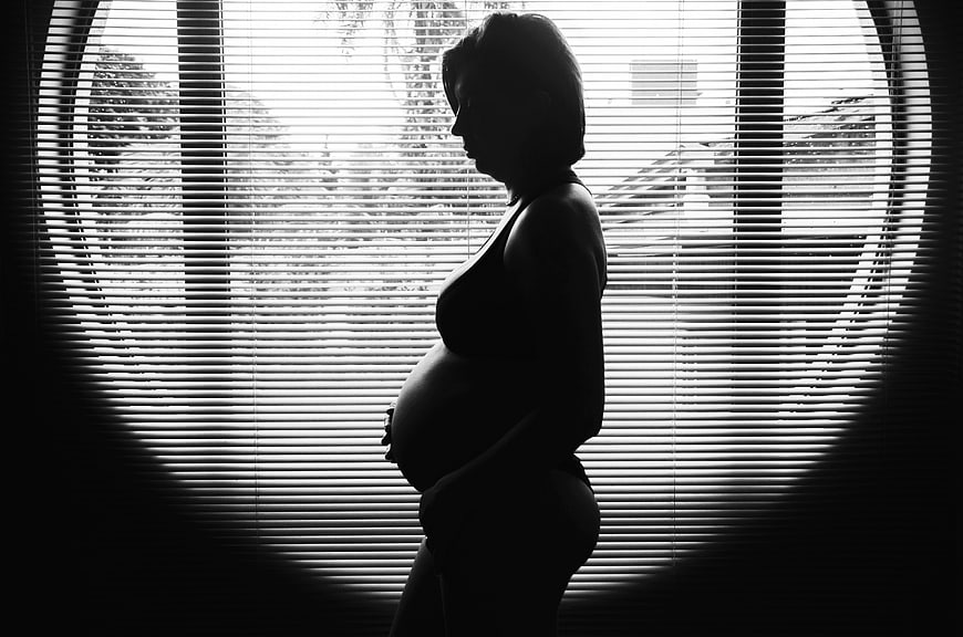 Silhouette of pregnant woman standing by window blinds