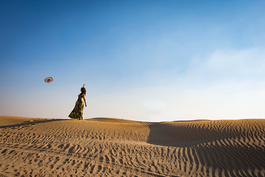 Woman Throwing Hat in Desert Dunes Under Blue Sky