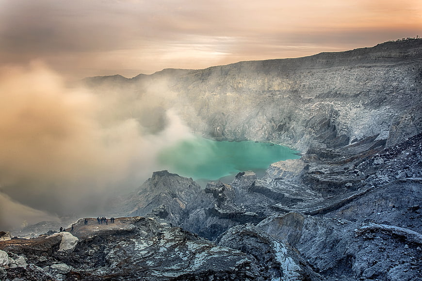 Turquoise volcanic crater lake with steam and hikers at sunrise