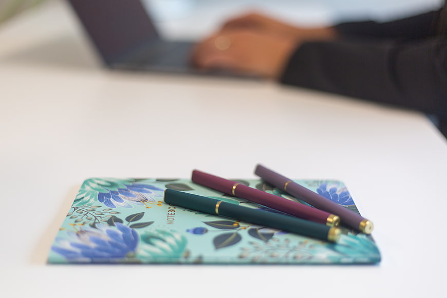 Floral Notebook and Pens on White Desk with Blurred Laptop