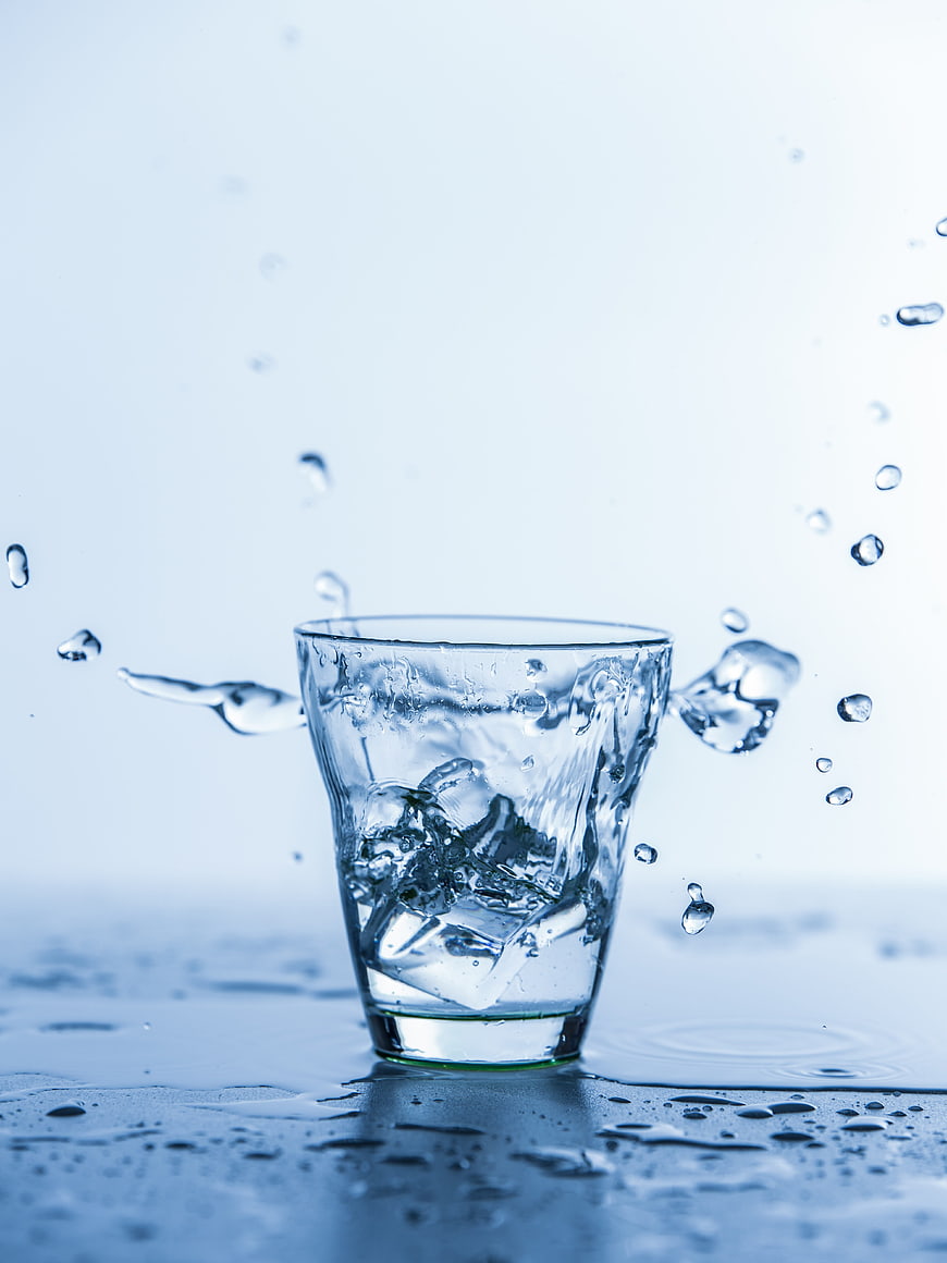 Glass of Ice Water with Splashing Droplets on Blue Background
