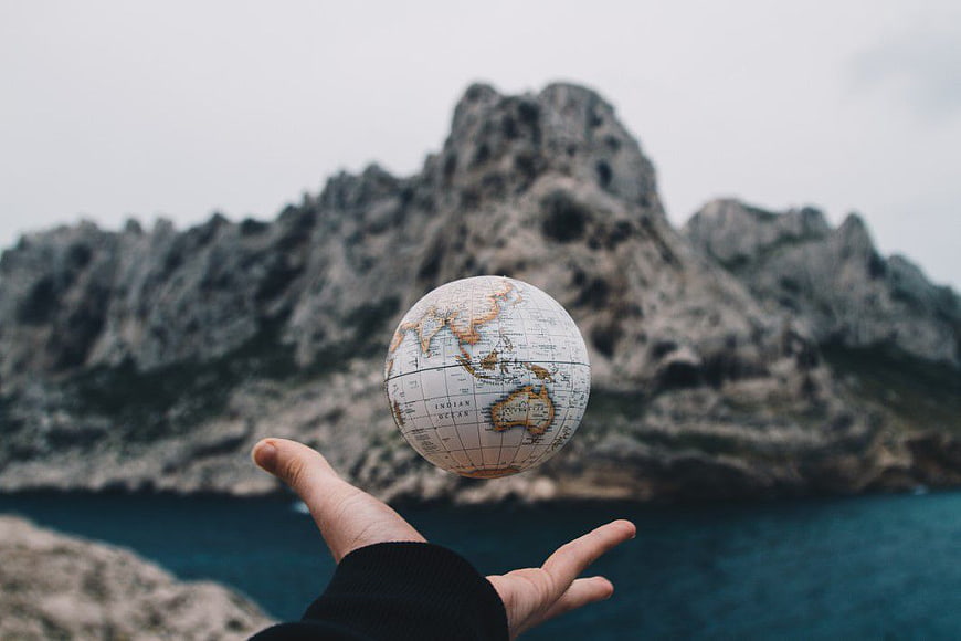 Hand Tossing Miniature Globe Against Coastal Mountain Landscape