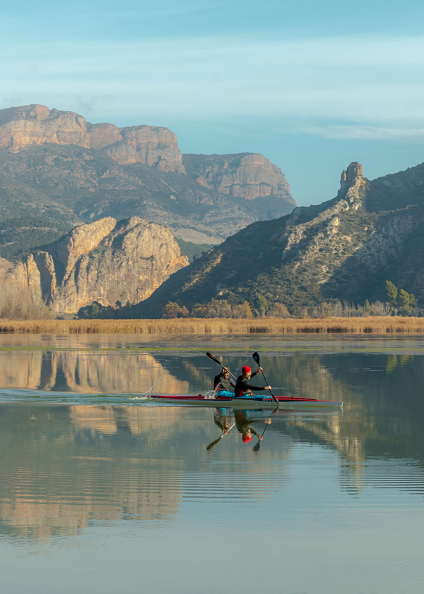 Two kayakers paddling on calm lake with mountain reflections