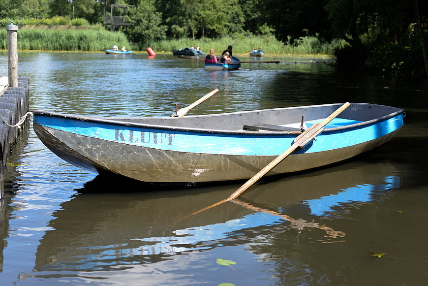 Blue wooden rowboat docked at a calm lake with people rowing in background