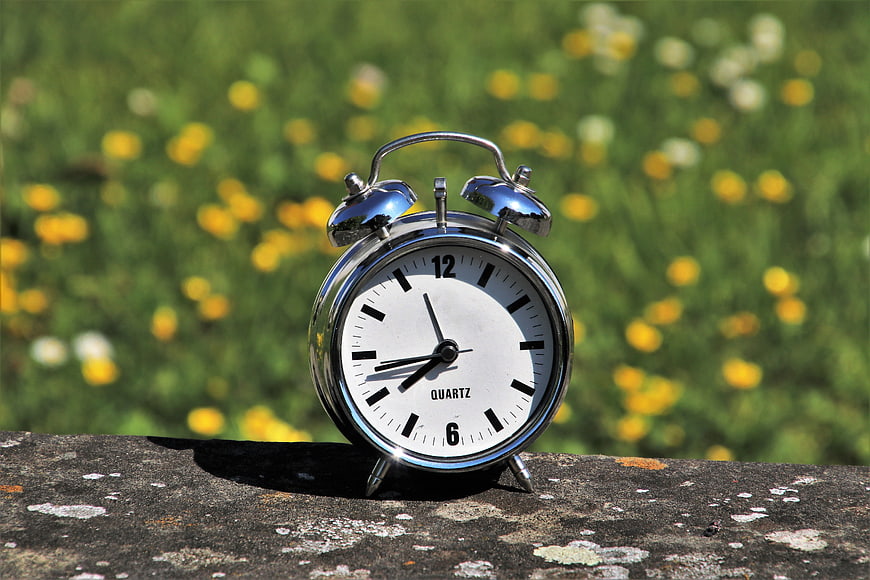 Silver alarm clock on stone wall with blurred yellow flowers