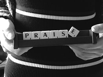 Hands holding Scrabble tiles spelling the word PRAISE in black and white