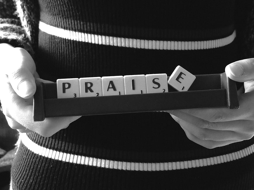 Hands holding Scrabble tiles spelling the word PRAISE in black and white