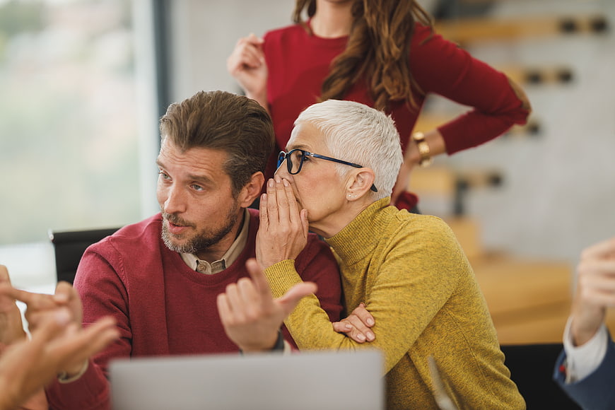 Senior woman whispering secret to male colleague in office meeting
