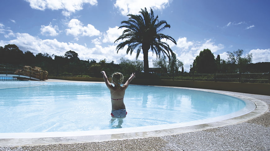 Child in pool with arms raised under palm tree