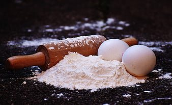 Baking ingredients with rolling pin, eggs, and flour on dark surface