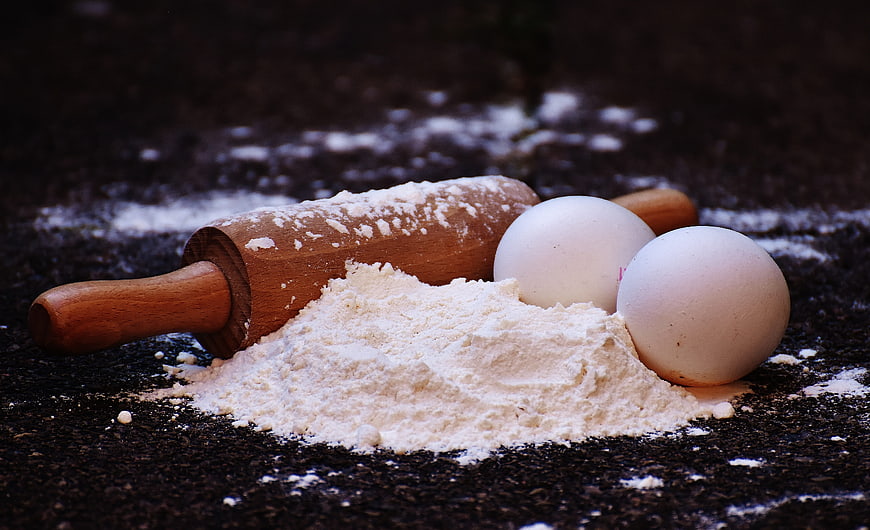 Baking ingredients with rolling pin, eggs, and flour on dark surface