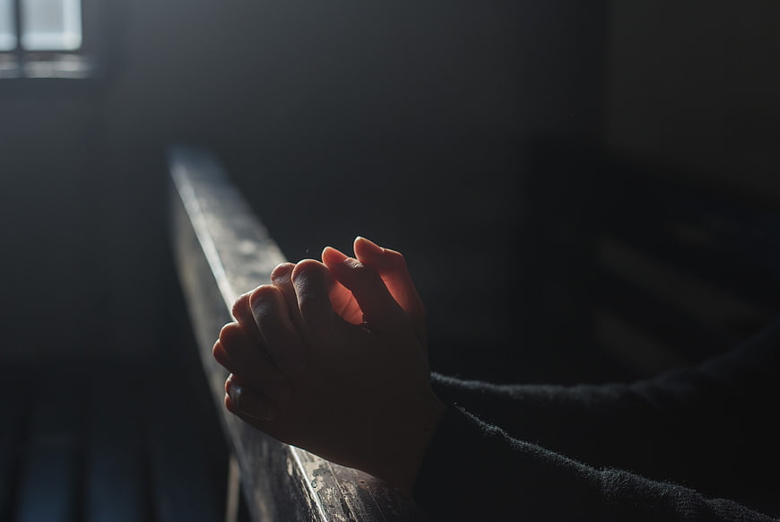 Close-up of hands clasped in prayer in a dimly lit church pew