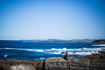 Man sitting on rocks overlooking blue ocean waves and distant mountains