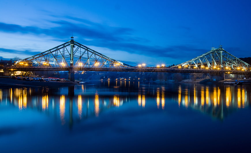 Illuminated Steel Truss Bridge At Dusk With Water Reflections