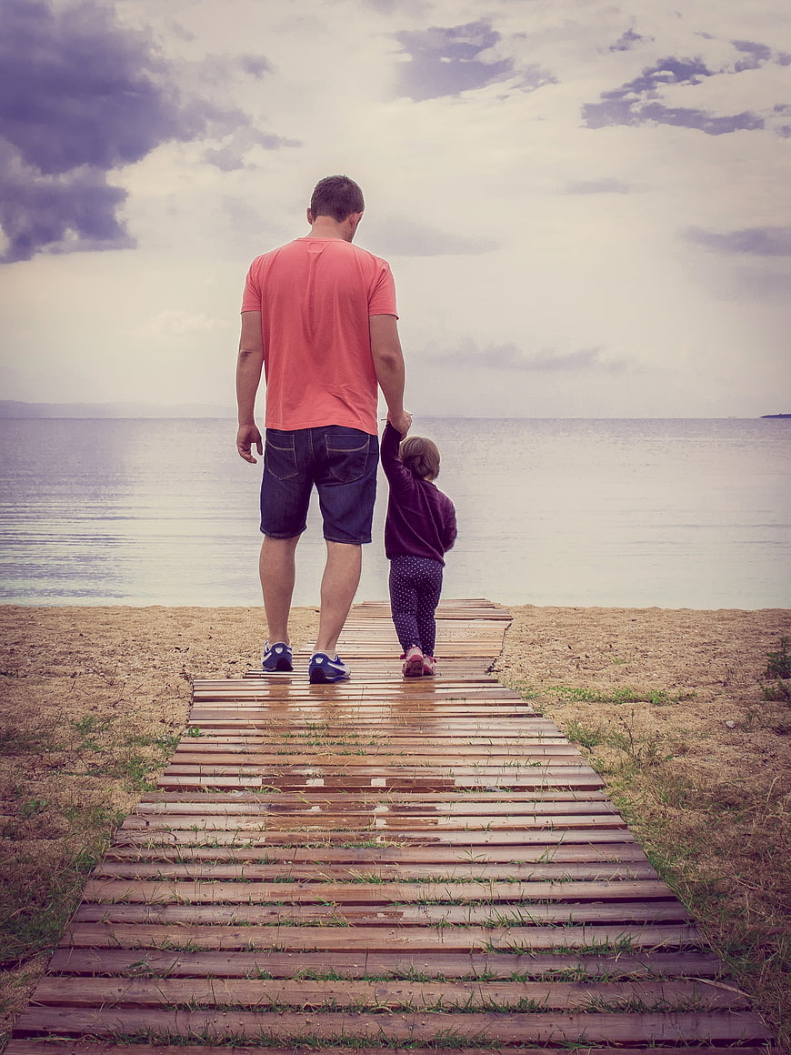 Father and daughter holding hands walking on wooden boardwalk by sea