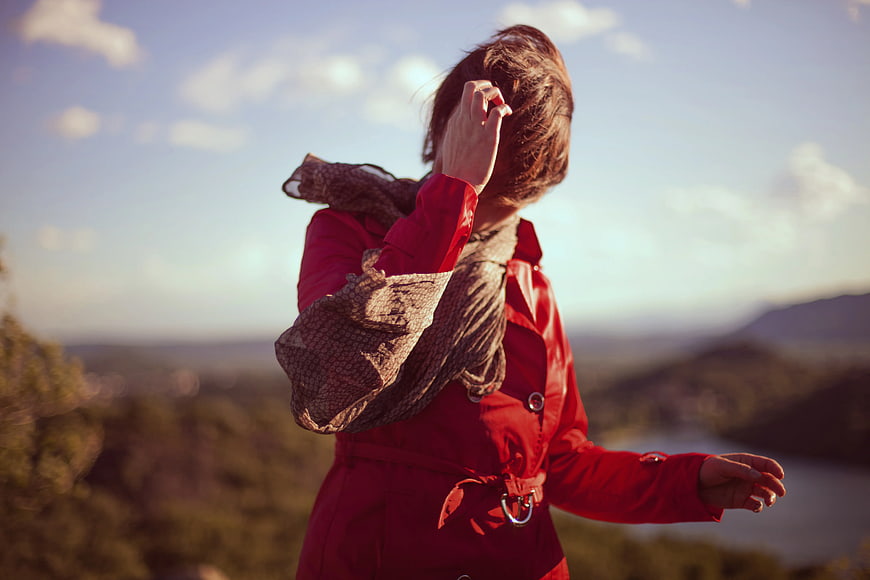 Woman in Red Coat Touching Hair in Scenic Landscape