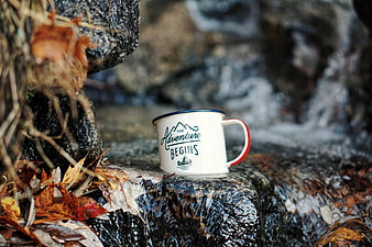 White enamel mug with adventure text on wet rock by stream