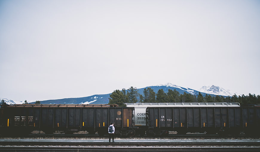 Man Standing by Freight Train Tracks with Snowy Mountains
