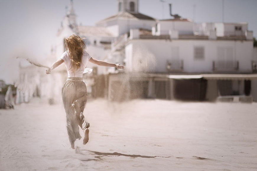 Woman Running Barefoot on Sandy Beach with White Buildings
