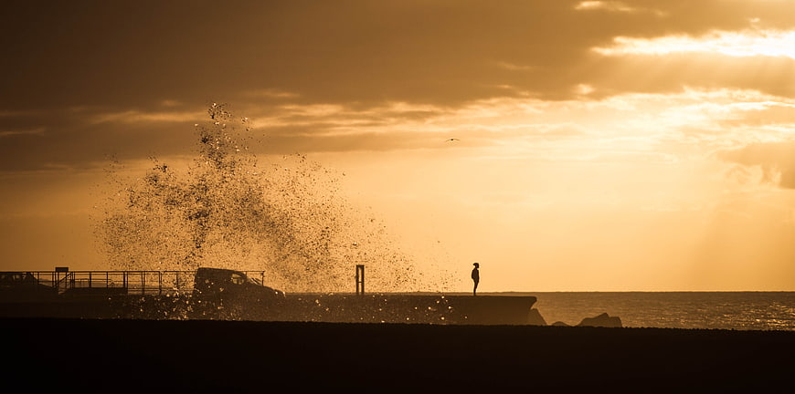 Silhouette of person on pier with crashing wave at sunset