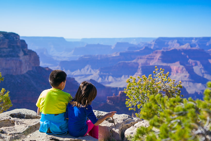 Two children sitting on a cliff edge overlooking a vast canyon
