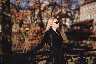 Stylish blonde woman in black coat and sunglasses on autumn bridge