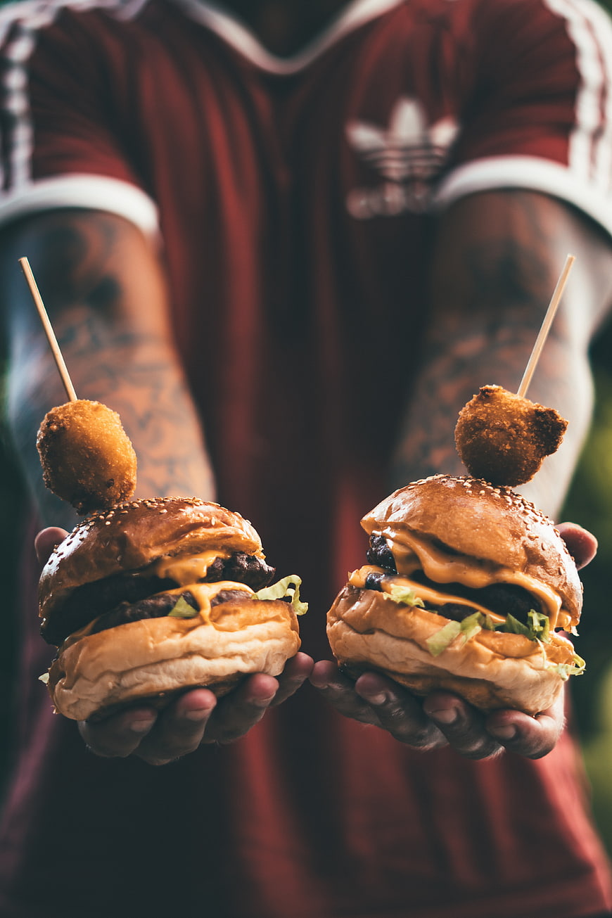 Man holding two gourmet burgers with fried toppings