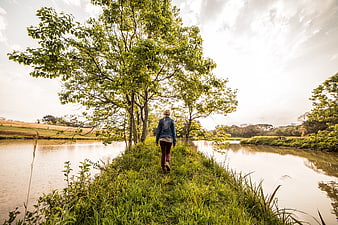 Woman Walking on Riverbank Path Under Green Trees