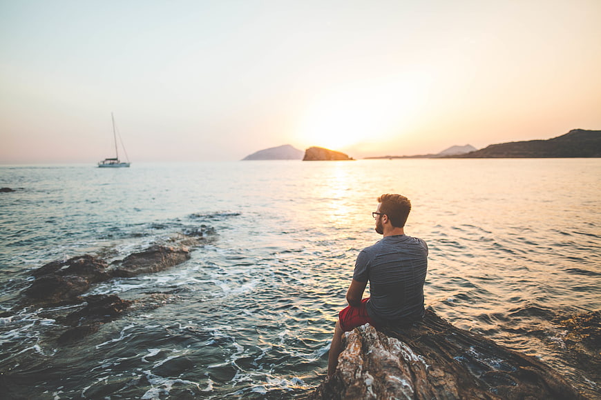 Man sitting on rocks watching sunset over ocean with sailboat
