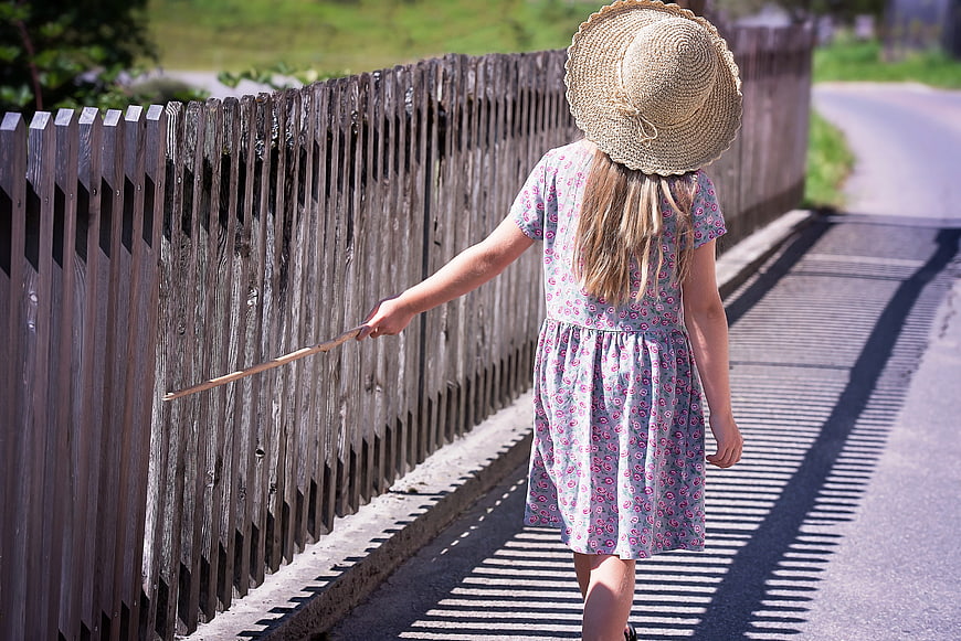 Little girl in straw hat walking along wooden fence