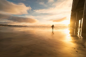 Surfer walking on wet beach at sunset under pier