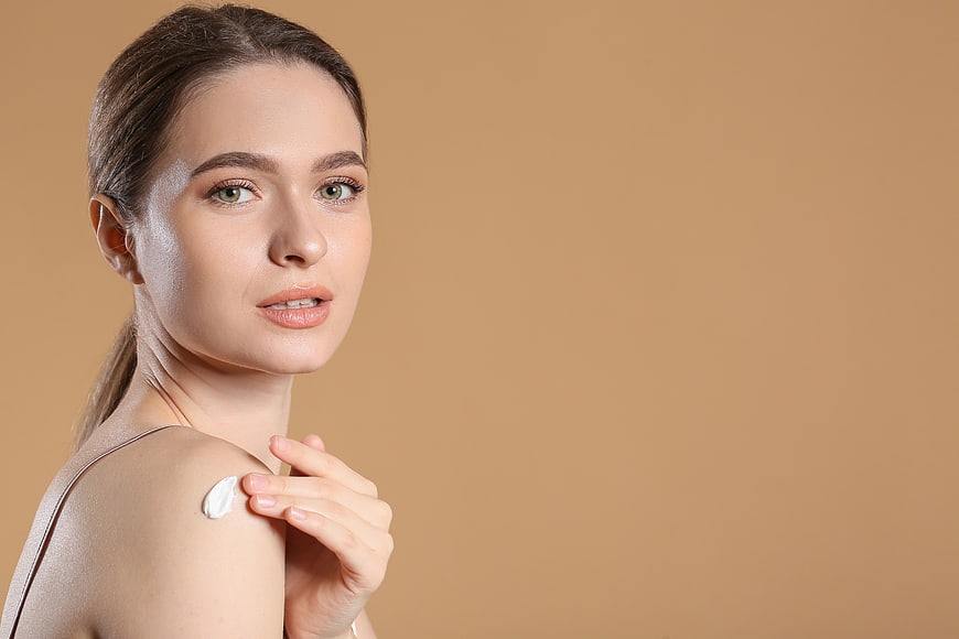 Woman applying moisturizer cream on shoulder against beige background