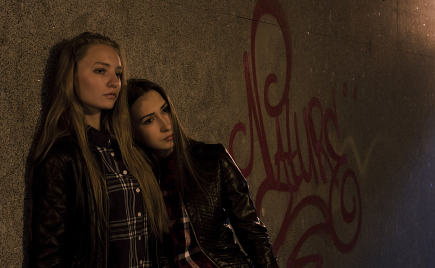Two women in leather jackets leaning against graffiti wall at night