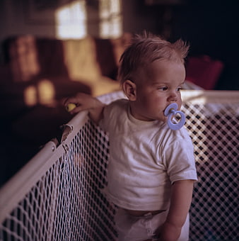 Toddler with pacifier standing in white mesh playpen