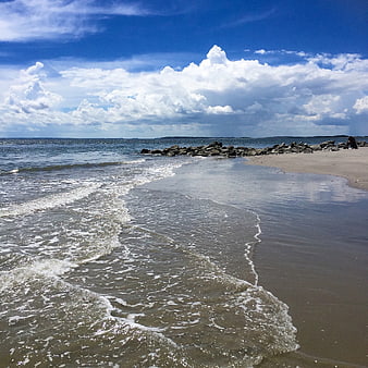 Sandy Beach with Gentle Waves and Rocky Jetty Under Blue Sky