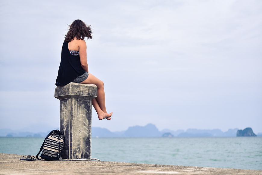Woman sitting on pier post overlooking calm sea and distant mountains