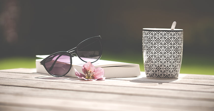 Sunglasses, book, and patterned mug on wooden table outdoors