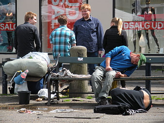 Homeless men sleeping on street bench with passersby