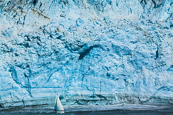 Small sailboat in front of massive blue glacier wall