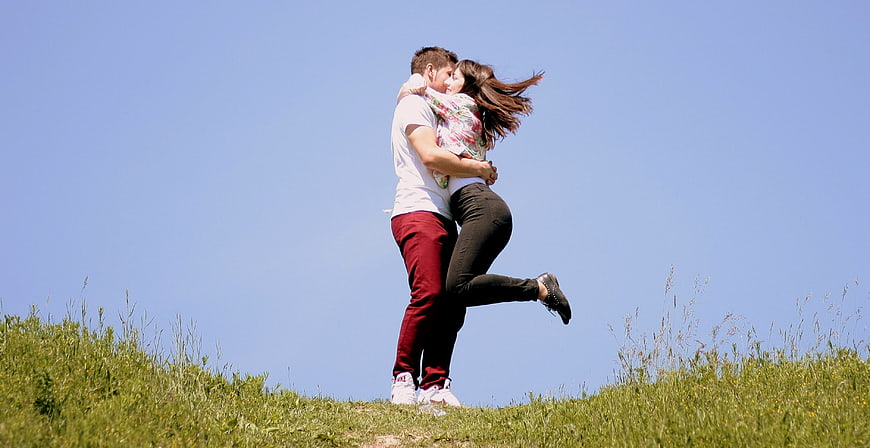 Romantic couple embracing and kissing on a grassy hill under a clear blue sky