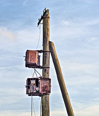 Rusty electrical boxes on weathered wooden utility pole against sky