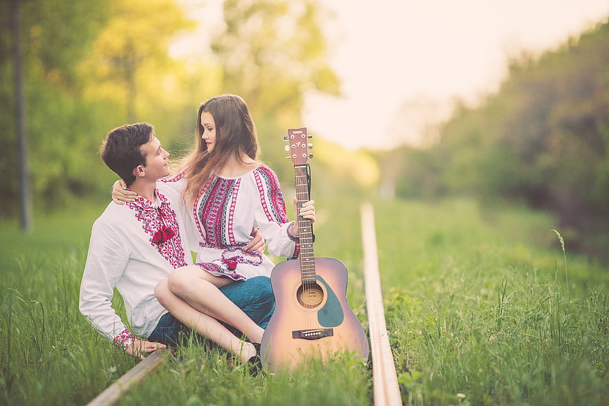 Young couple in traditional embroidered shirts sitting on railway tracks with guitar