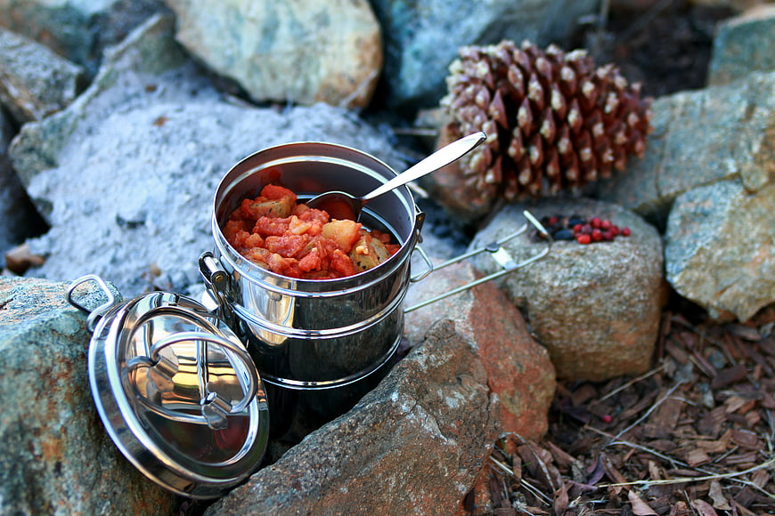 Camping stew in stainless steel pot with pine cone and nuts on rocks